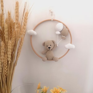 Peut inclure: Mobile de chambre d'enfant fait à la main, avec un ours en peluche, deux ballons et deux nuages en crochet, suspendus dans un cercle en bois. L'ours est marron clair, les ballons sont blancs et beiges, et les nuages sont blancs.