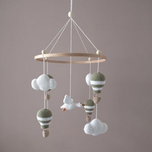 Peut inclure: Un mobile en crochet blanc et vert avec des nuages, des ballons à air chaud et un oiseau suspendu à un anneau en bois.