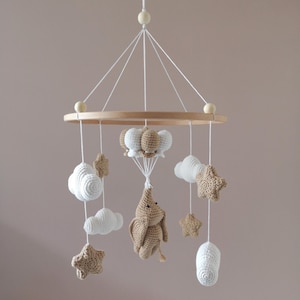 Op de afbeelding: Handgemaakte baby mobile met een houten ring en hangende gehaakte elementen. De mobile bevat witte wolken, beige sterren en een beige konijn dat aan witte ballonnen hangt. Het ontwerp is neutraal en geschikt voor een kinderkamer.