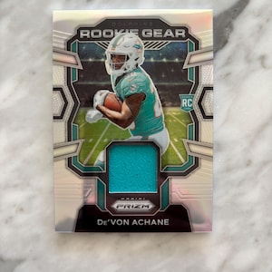 May include: A De'Von Achane rookie card from the Panini Prizm series. The card features a player in a Miami Dolphins uniform, with the words "ROOKIE GEAR" at the top. The card has a teal jersey insert.