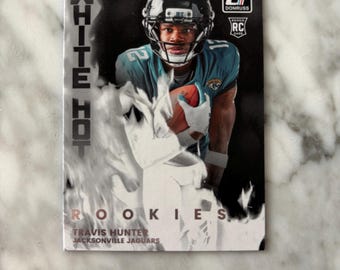 May include: A Donruss football trading card featuring Travis Hunter of the Jacksonville Jaguars. The card has a photo of Hunter in his team uniform, with the words "WHITE HOT" and "ROOKIES" in white text. The card also has the Donruss logo.