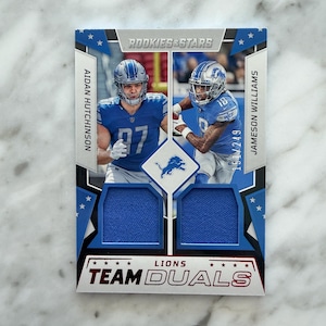 May include: A Rookies & Stars Team Duals trading card featuring Detroit Lions players Aidan Hutchinson and Jameson Williams. The card displays two blue jersey swatches, the Lions logo, and the text 151/249, set against a blue and silver frame.