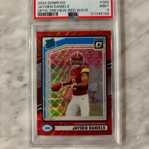 May include: A graded football card featuring Jayden Daniels in a Washington Commanders uniform. The card has a red Optic Preview-Red Wave design with the text "2024 Donruss Jayden Daniels" and "Optic" branding. The card is graded as Mint 9.