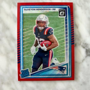 May include: A red and blue TreVeyon Henderson football card. The card features a player in a Patriots uniform holding a football. The card has the text "Rated Rookie" and "Optic".