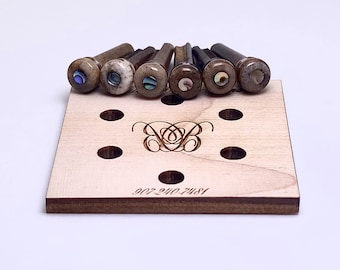May include: A collection of guitar bridge pins with abalone shell inlays, displayed on a light wooden holder. The pins are various shades of brown and gray. The holder has a decorative design and the phone number 907-240-7481.