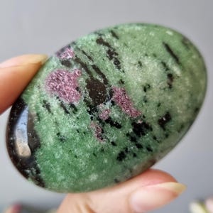 May include: Oval-shaped gemstone with a polished surface. The stone has a green base colour with black and pink inclusions. The gemstone is held between fingers, showcasing its smooth texture and natural patterns. A unique, natural stone.