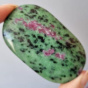 May include: A smooth, oval gemstone with a green base, black speckles, and patches of pink. The stone is held in a hand, highlighting its polished surface and natural colour variations. The stone's shape is approximately 7 cm long.