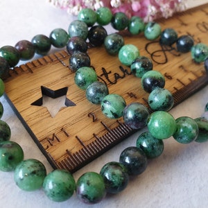 May include: A green and black gemstone bracelet with round beads. The bracelet is laid out on a wooden ruler with a star design.