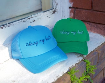 May include: Two trucker hats with a mesh back. The hats are blue and green and both have the text "#doing my best" embroidered on the front.