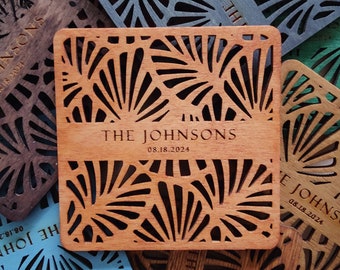 May include: A collection of square wooden coasters in various colors, including brown, blue, and green. Each coaster features a laser-cut geometric design and the text "THE JOHNSONS 08.18.2024". The coasters are arranged on a decorative plate.