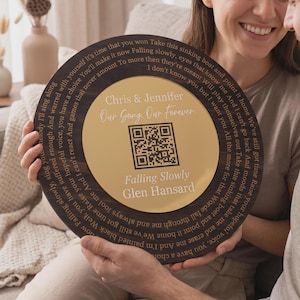 May include: A round wooden plaque with a gold center is held by a couple. The plaque has engraved text and a QR code. The text includes the names "Chris & Jennifer" and the song title "Falling Slowly" by Glen Hansard.