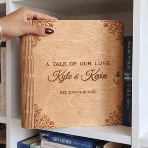 Può includere: Un album fotografico quadrato in legno con finitura marrone chiaro. La copertina presenta decorazioni floreali incise al laser negli angoli e il testo A TALE OF OUR LOVE, Kyle & Kevin, e EST. AUGUST 16. 2025. Un piccolo ciondolo a cuore è fissato sul lato destro.