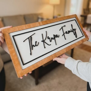 Puede incluir: Un letrero enmarcado de madera con las palabras "The Krupa Family" en escritura negra. El letrero tiene un fondo blanco y un borde negro. El marco es de color marrón claro.