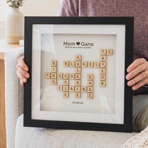 May include: A framed artwork with the words "Mom Gana" and Scrabble tiles spelling out names and words. The black frame surrounds a white mat, and the text "Built by love, letter by letter" is also included. The score is 85.