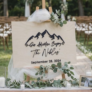 May include: A wooden sign on an easel with the text "Our Adventure Begins" and "The Nobles" with the date September 28, 2024. The sign features a mountain range design. The sign is decorated with greenery and white flowers.
