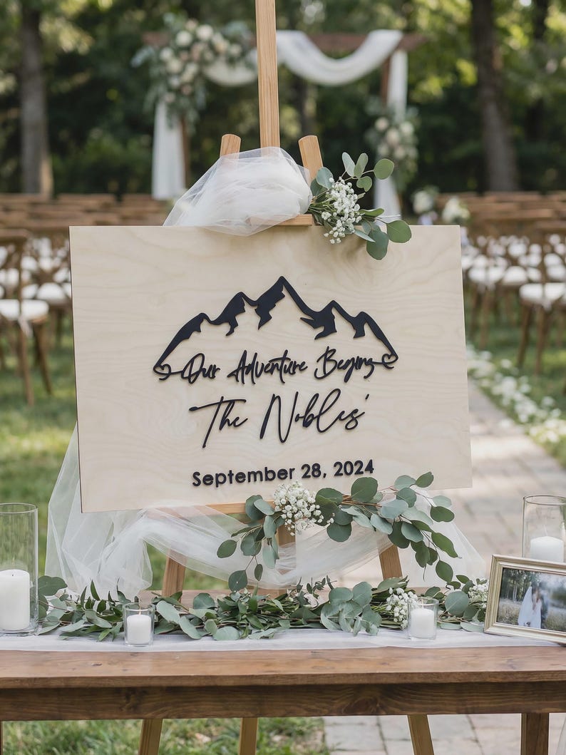 May include: A wooden sign on an easel with the text "Our Adventure Begins" and "The Nobles" with the date September 28, 2024. The sign features a mountain range design. The sign is decorated with greenery and white flowers.