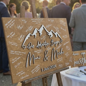 May include: A wooden sign on an easel, featuring the words "Our Adventure Begins", the names "Moni & Michi", and the date "1. Juni 2024". The sign has a mountain design and is covered in handwritten messages.