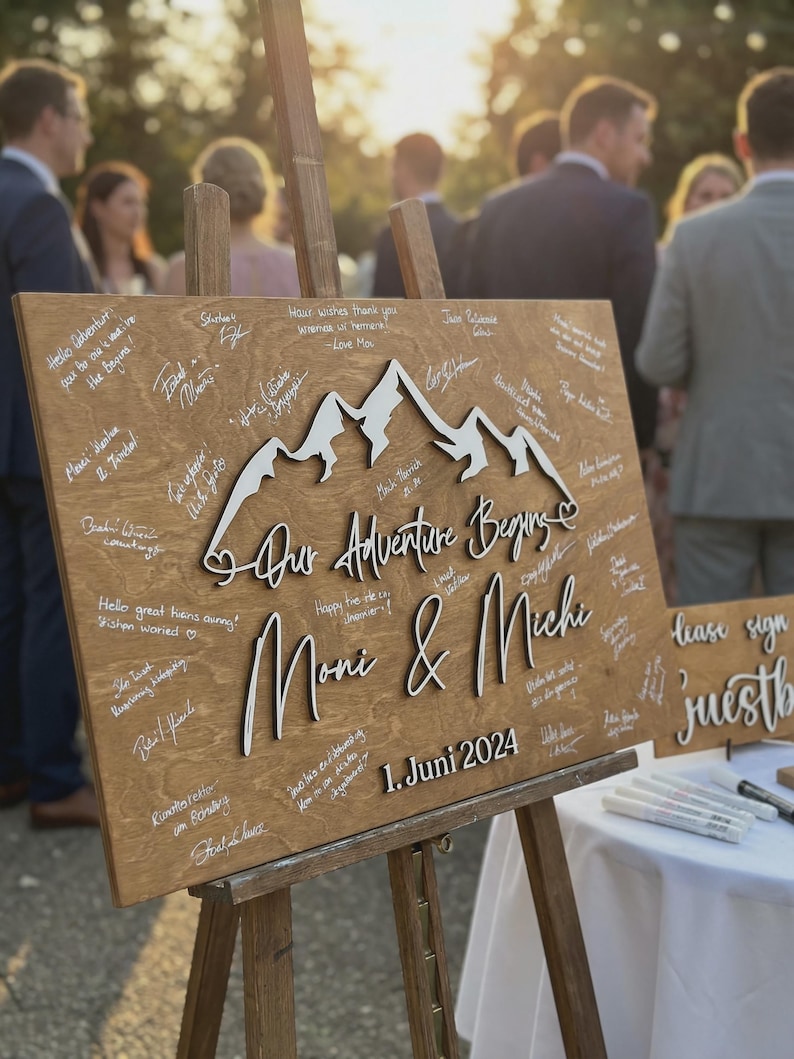May include: A wooden sign on an easel, featuring the words "Our Adventure Begins", the names "Moni & Michi", and the date "1. Juni 2024". The sign has a mountain design and is covered in handwritten messages.