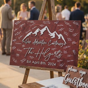 May include: A wooden wedding sign with the text "Our Adventure Continues" and "The Hilgerts" with the date "Est April 8th, 2024". The sign features a mountain range design and is propped on a wooden easel. Guests' signatures are visible.