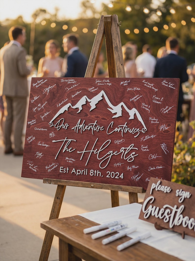 May include: A wooden wedding sign with the text "Our Adventure Continues" and "The Hilgerts" with the date "Est April 8th, 2024". The sign features a mountain range design and is propped on a wooden easel. Guests' signatures are visible.