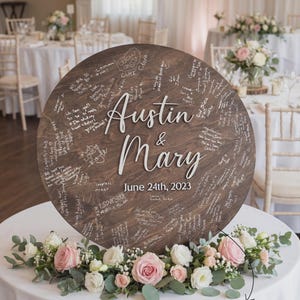 Large round wooden wedding guestbook sign displayed upright on a white table, personalized with the couple’s names and wedding date in white lettering. The surface is filled with handwritten guest messages. Floral arrangements surround the base, with a decorated reception room in the background.