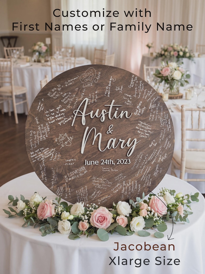 Large round wooden wedding guestbook sign displayed upright on a white table, personalized with the couple’s names and wedding date in white lettering. The surface is filled with handwritten guest messages. Floral arrangements surround the base, with a decorated reception room in the background.