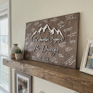 Rustic personalized wooden wedding guest book alternative wall sign with mountain design, custom wedding guestbook with names and date displayed on home shelf with guest signatures.