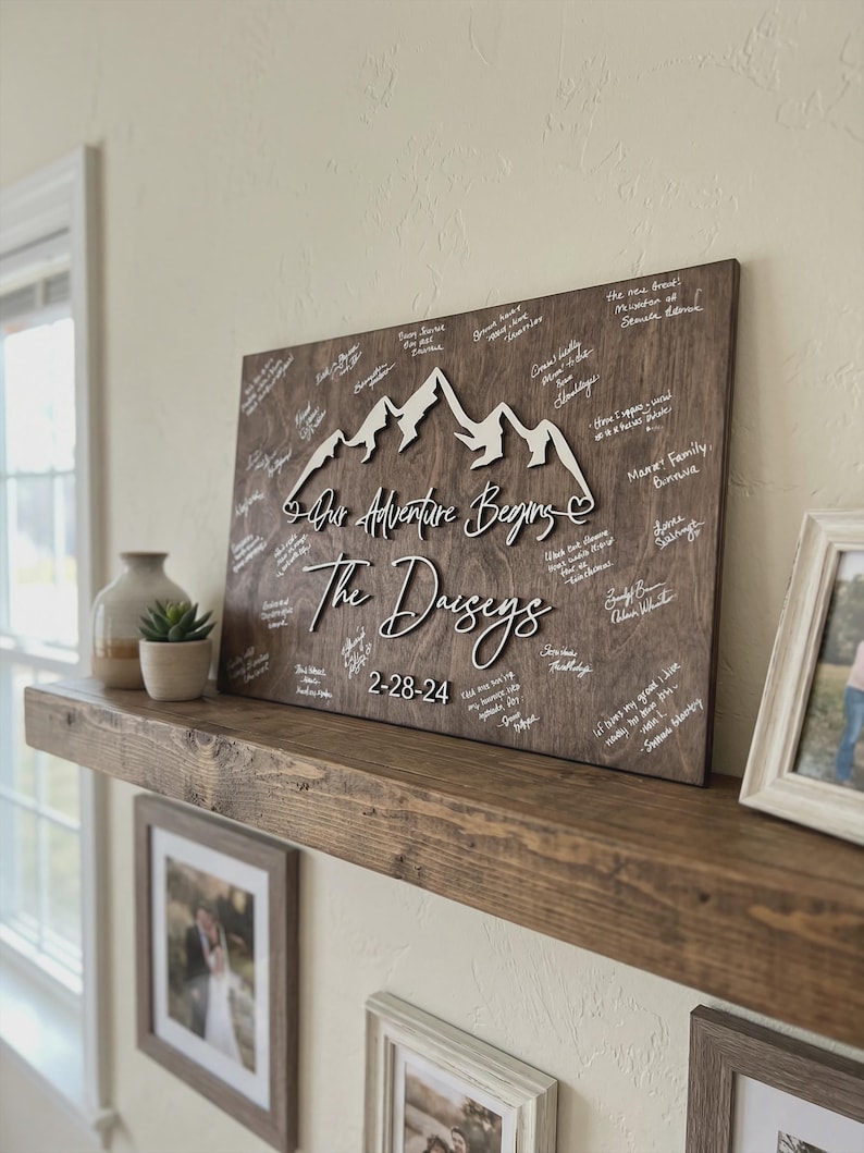 Rustic personalized wooden wedding guest book alternative wall sign with mountain design, custom wedding guestbook with names and date displayed on home shelf with guest signatures.