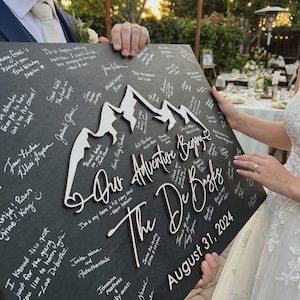 May include: A black wooden sign featuring white mountain cutouts and the text "Our Adventure Begins" and "The De Busks", dated August 31, 2024. The sign is covered in handwritten messages.