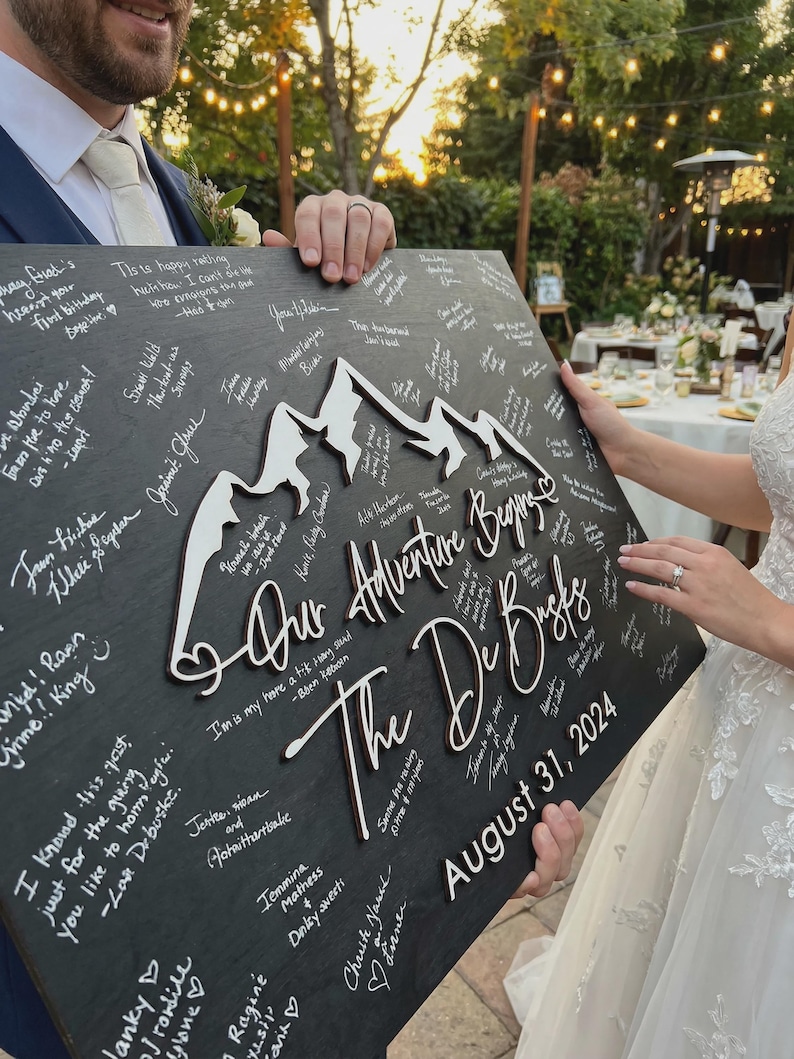 May include: A black wooden sign featuring white mountain cutouts and the text "Our Adventure Begins" and "The De Busks", dated August 31, 2024. The sign is covered in handwritten messages.
