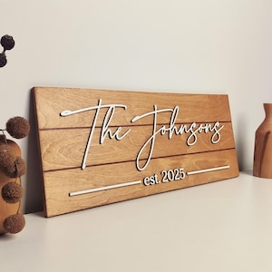 Op de afbeelding: Houten familienaambord met de tekst "The Johnsons" in een witte, cursieve letter. Het bord is gemaakt van lichtbruin hout met horizontale lijnen en de tekst "est. 2025" in een witte, schreefloze letter.
