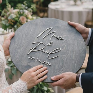 May include: A circular, grey wooden sign featuring the names "Maxi & Paul" and the date "Juli 13, 2024" in white script. The sign is held by two people, possibly at a wedding. The background includes foliage and a table.