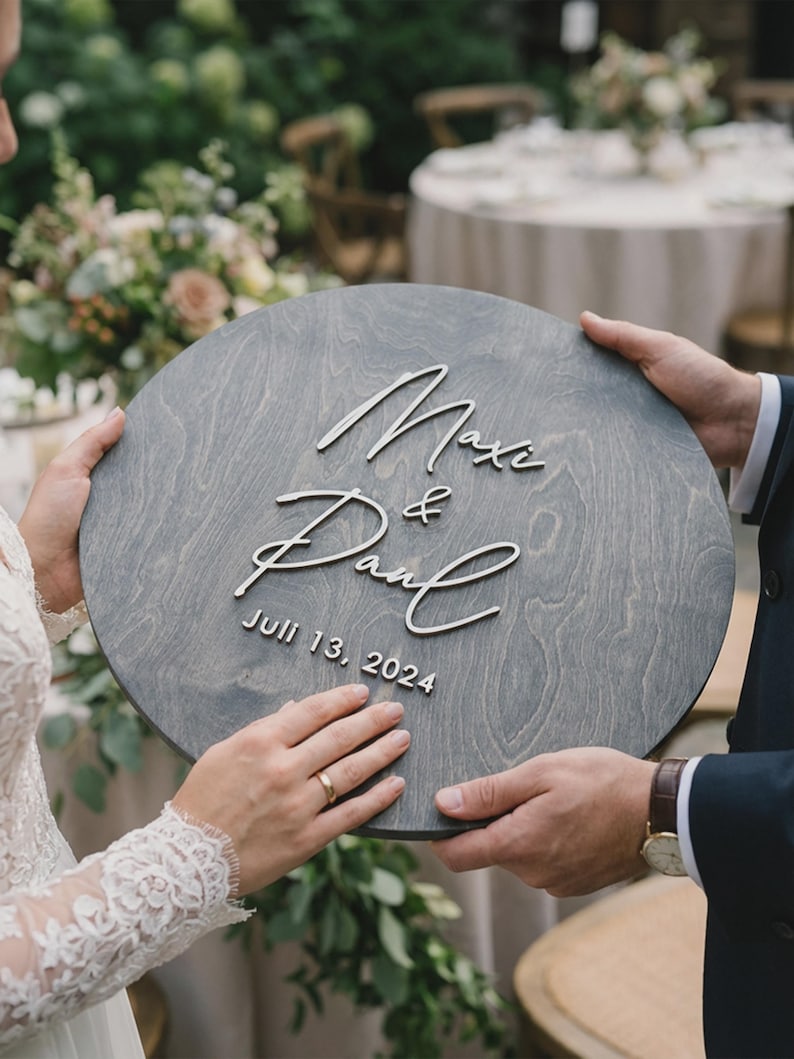 May include: A circular, grey wooden sign featuring the names "Maxi & Paul" and the date "Juli 13, 2024" in white script. The sign is held by two people, possibly at a wedding. The background includes foliage and a table.