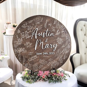 May include: A round wooden wedding sign with the names "Austin & Mary" in white script, and the date "June 24th, 2023". The sign is covered in handwritten messages and rests on a stand with floral arrangements.