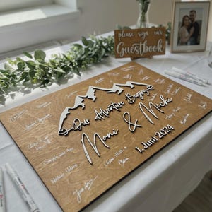 May include: A wooden wedding guest book with a mountain design and the words "Our Adventure Begins Moni & Michi 1. Juni 2024". The wooden board is light brown with white signatures and a white mountain range.