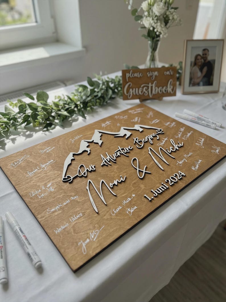 May include: A wooden wedding guest book with a mountain design and the words "Our Adventure Begins Moni & Michi 1. Juni 2024". The wooden board is light brown with white signatures and a white mountain range.