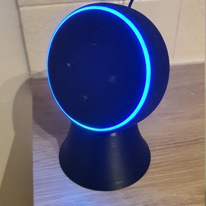 May include: A black smart speaker with a blue light ring around the top. The speaker has volume controls and a microphone button. The speaker is sitting on a wooden surface and is plugged into an outlet.