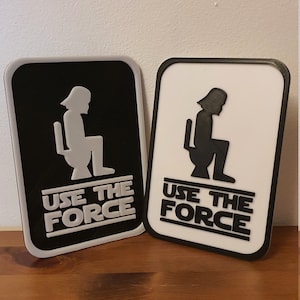 May include: Two black and white signs with a silhouette of a person sitting on a toilet. The signs say "Use the Force" in white letters.