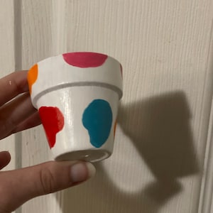May include: A small, white ceramic flower pot with a gray rim and colorful, abstract polka dots in red, orange, pink, and blue. The pot is held against a white background.