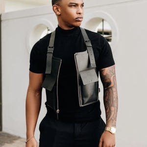 Puede incluir: Arnés de pecho de cuero negro con cremallera y dos bolsillos delanteros. El arnés tiene correas ajustables y un diseño minimalista. El arnés se lleva sobre una camisa negra.