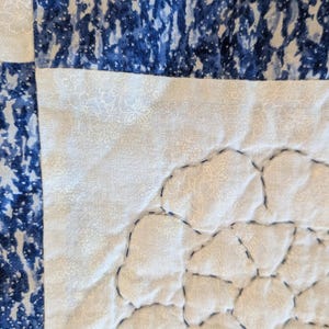 May include: Close-up of a quilted fabric with a patchwork design. The quilt features sections of white fabric with a subtle floral pattern, and sections of blue fabric with a mottled, watercolor-like effect. Dark blue stitching outlines the shapes.