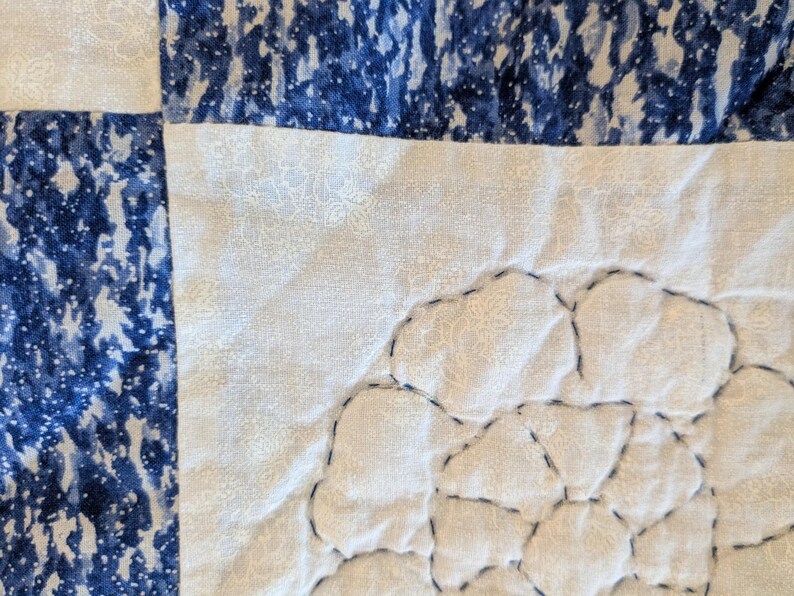May include: Close-up of a quilted fabric with a patchwork design. The quilt features sections of white fabric with a subtle floral pattern, and sections of blue fabric with a mottled, watercolor-like effect. Dark blue stitching outlines the shapes.