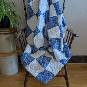 May include: A blue and white quilt with a floral pattern is draped over a wooden chair. The quilt features a checkerboard design with alternating squares of blue speckled fabric and white squares with embroidered floral motifs. The chair is made of dark wood.