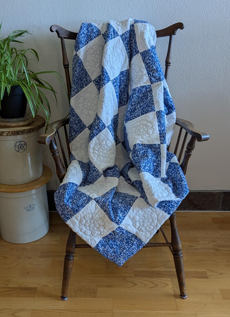 May include: A blue and white quilt with a floral pattern is draped over a wooden chair. The quilt features a checkerboard design with alternating squares of blue speckled fabric and white squares with embroidered floral motifs. The chair is made of dark wood.