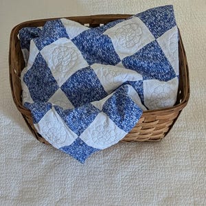 May include: A blue and white checkered quilt with floral embroidery is nestled inside a brown wicker basket. The quilt's design features alternating squares of solid white and blue speckled fabric. The basket sits on a white textured surface.