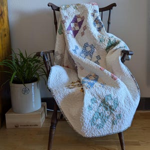 May include: A vintage quilt draped over a wooden chair. The quilt features a white background with various colorful patchwork designs, including diamonds and floral patterns. The chair is dark brown, and a potted plant sits nearby.