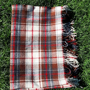 May include: A folded plaid blanket with a brown, white, red, and gray pattern. The blanket has a fringed edge with red, white, and black threads. The blanket is lying on a bed of green grass.