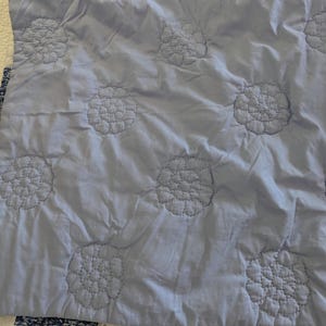 May include: A light blue quilt with a floral pattern. The quilt features large, embroidered flower designs evenly spaced across the surface. The stitching is a darker shade of blue, creating a subtle contrast. The quilt is folded, showing a portion of a patterned border.