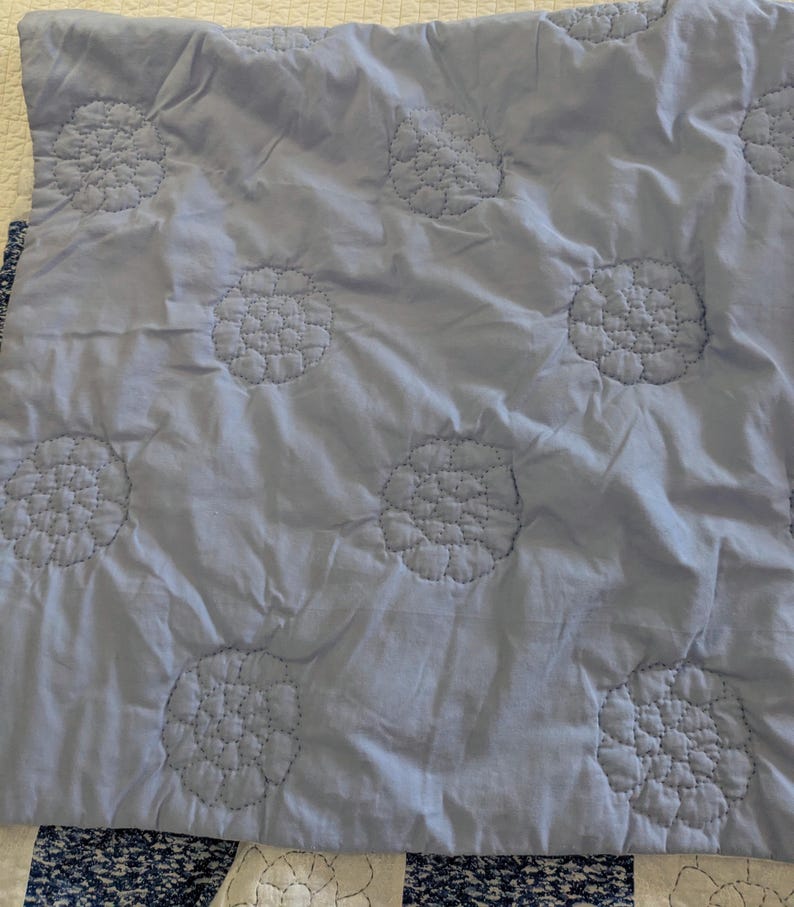 May include: A light blue quilt with a floral pattern. The quilt features large, embroidered flower designs evenly spaced across the surface. The stitching is a darker shade of blue, creating a subtle contrast. The quilt is folded, showing a portion of a patterned border.