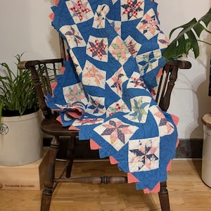 May include: A vintage quilt draped over a wooden chair. The quilt features a blue background with star-shaped patterns in various colors, including red, pink, and white. The edges are trimmed with a scalloped coral border, adding a decorative touch.